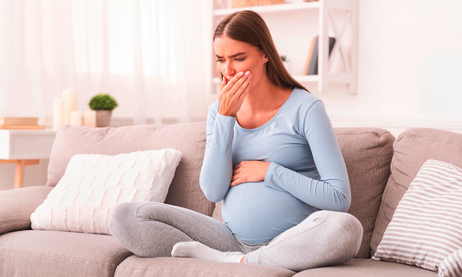 Eine schwangere Frau sitzt erkältet auf dem Sofa und hält sich die Hand vor dem Mund