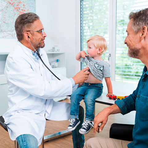 Ein Arzt hört einen kleinen Jungen an der Brust ab, sein Vater sitzt daneben