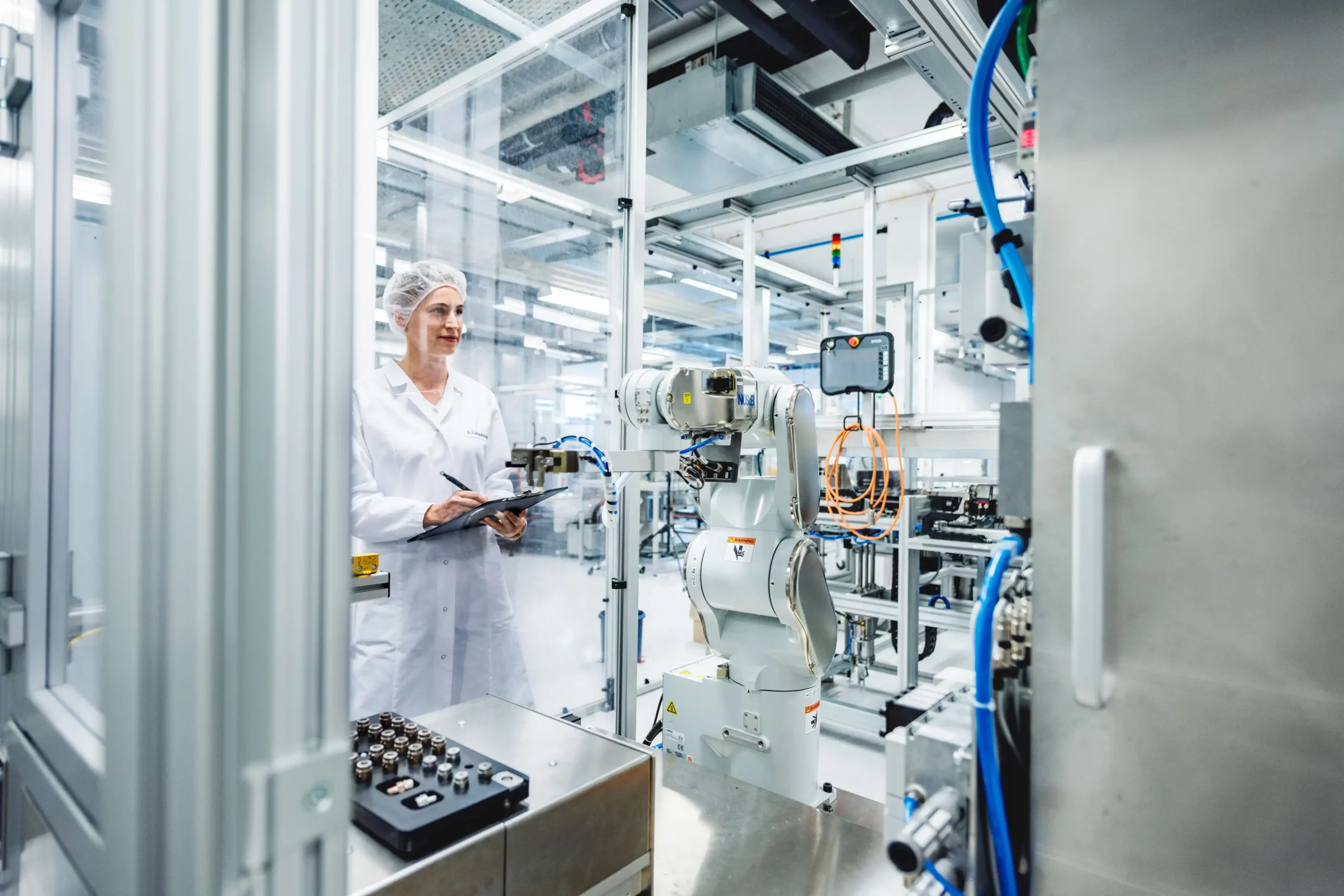 Female technician in a white lab coat and hairnet observing and taking notes as a robotic arm operates in a clean, high-tech manufacturing lab.