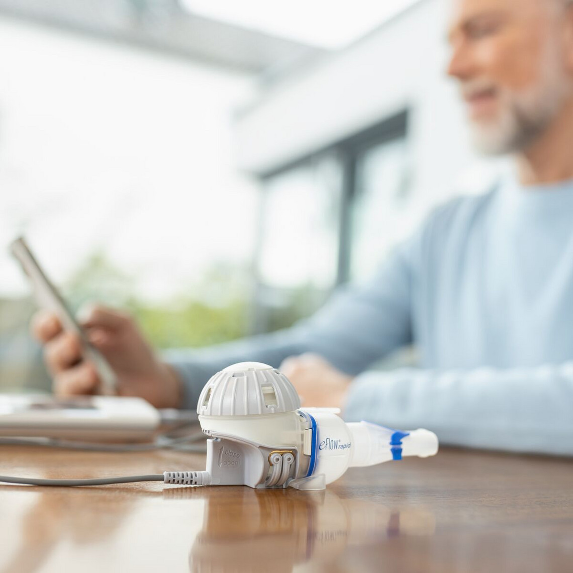 The eFlow rapid nebuliser system can be seen in the foreground, with an elderly man holding a smartphone in the background