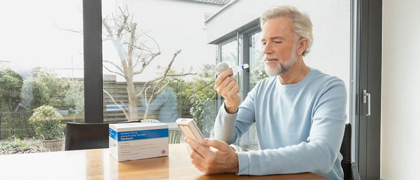 Woman sitting at a table using a nebuliser connected to an inhalation device while looking at a laptop, as a man stands nearby.