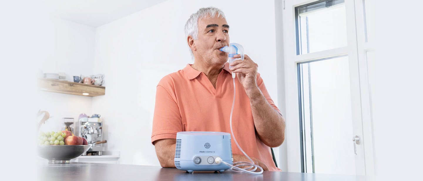 Ein älterer Mann mit grauen Haaren und einer korallfarbenen Polo-Shirts sitzt in einer modernen Küche und inhaliert mit dem PARI COMPACT2 Inhaliergerät. Er hält das Mundstück an den Mund, während das Gerät auf der dunklen Arbeitsplatte steht. Im Hintergrund sind eine Obstschale, eine Kaffeemaschine und ein Regal mit Küchenutensilien zu sehen.