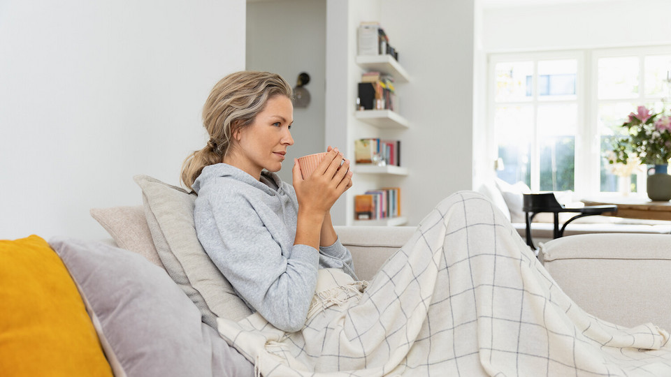 Junge, schwangere Frau sitzt mit Teetasse unter einer Decke auf der Couch
