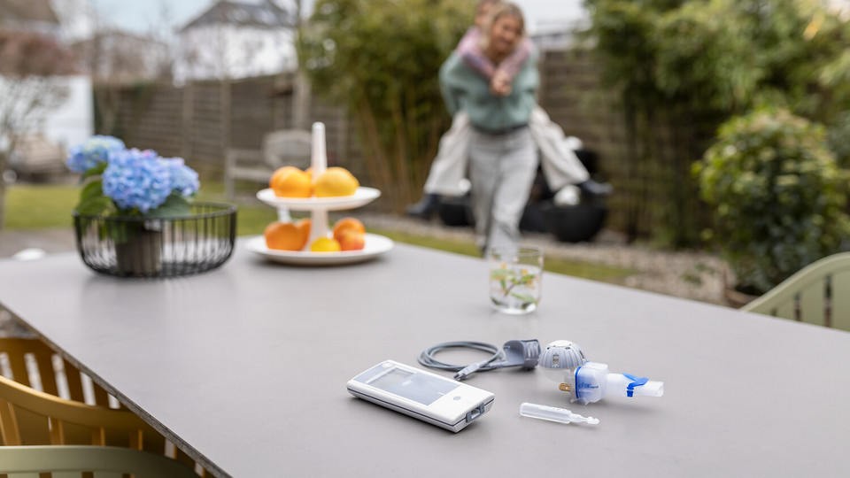 Interviews zum Umgang mit Keimen bei Kindern mit CF eFlow®rapid Inhalationssystem liegt auf dem Gartentisch, im Hintergrund spielt Mutter mit Kind