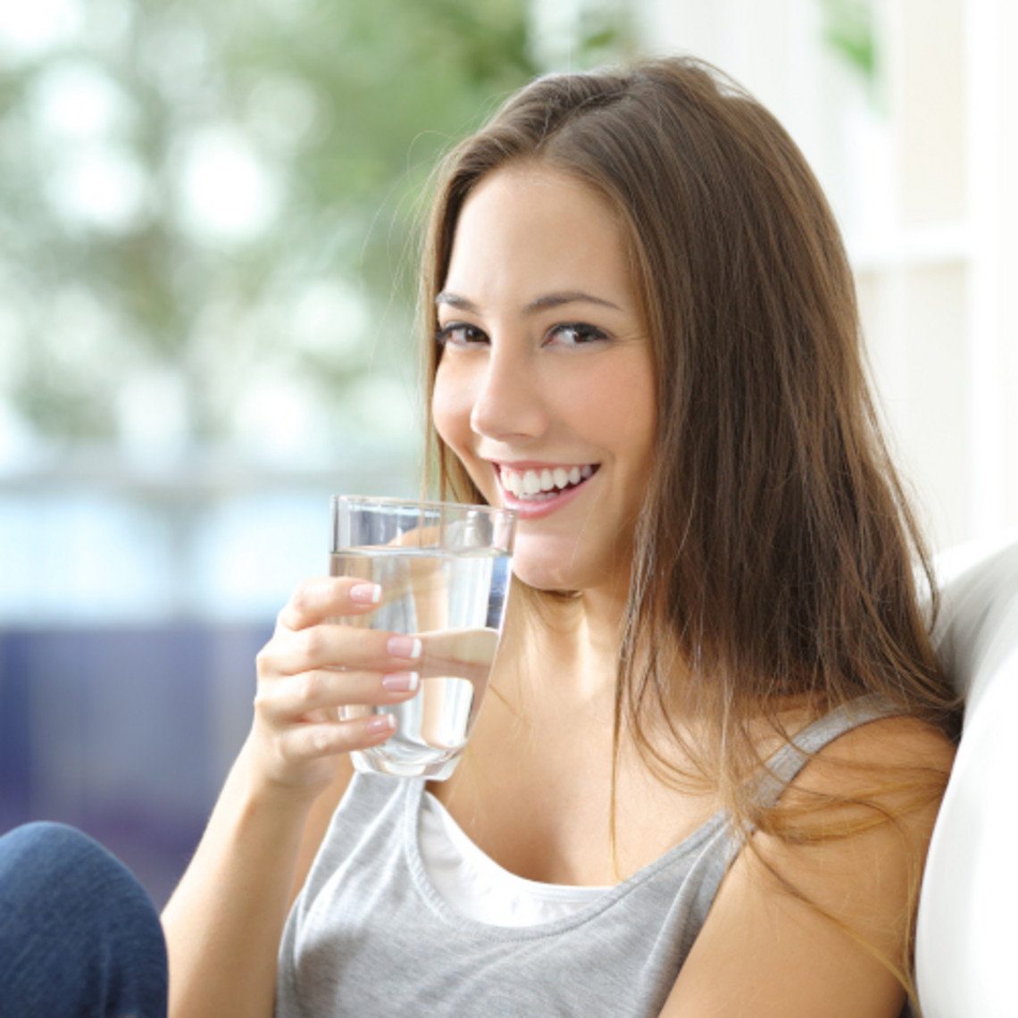 Nebenwirkungen von Kortisonsprays bei Asthma reduzieren: danach etwas trinken Lächelnde, junge Frau sitzt mit Glas Wasser in der Hand auf dem Sofa