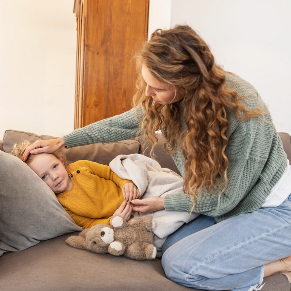 Mädchen liegt auf der Couch, Mutter sitzt daneben und hält die Hand auf die Stirn der Tochter