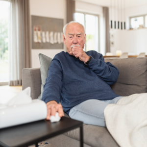 Älterer Mann sitzt halb liegend auf dem Sofa und hustet während er nach einem Taschentuch greift