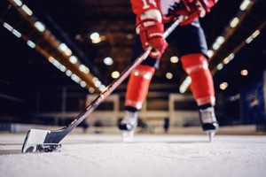 Nahaufnahme eines Hockeyschlägers mit Puk auf dem Eis, im Hintergrund sind Beine des Spielers zu erkennen