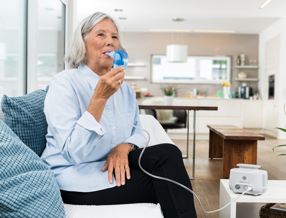 An elderly woman sits relaxed on a sofa in a living room. She uses a PARI LC SPRINT nebulizer connected via a tube to the PARI COMPACT2 compressor on the table.