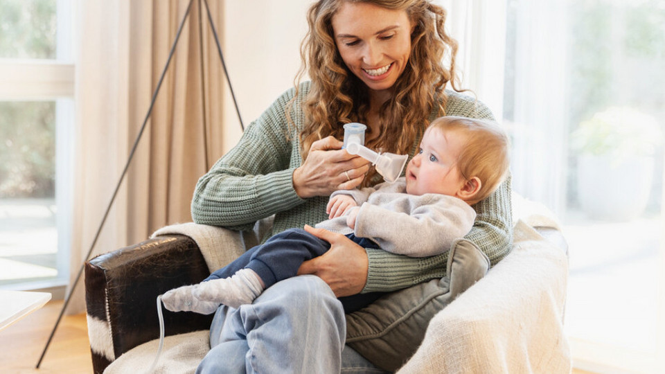 Junge Mutter sitzt mit Baby auf Sessel, Baby inhaliert mit Babywinkel und Babymaske