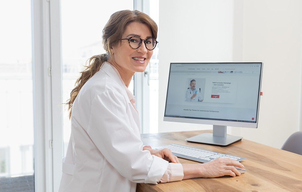 Person in weißem Kittel nutzt Computer mit medizinischem Login-Portal in heller Praxis mit Pflanzen und Sitzbereich.