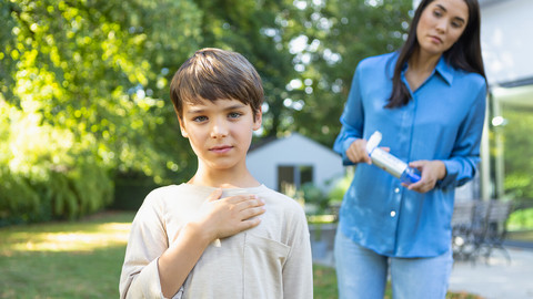 Asthmaanfall bei Kindern: Was Eltern tun sollten Kind hält sich die Hand auf Brust, Mutter steht mit VORTEX Inhalierhilfe und Asthmaspray im Hintergrund