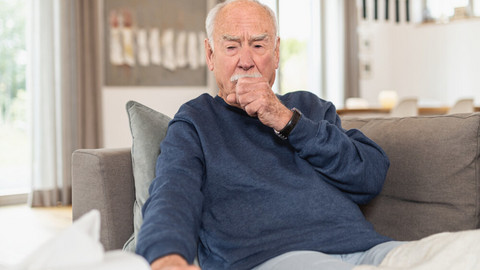 Älterer Mann sitzt hustend mit Faust vor dem Mund auf Sofa