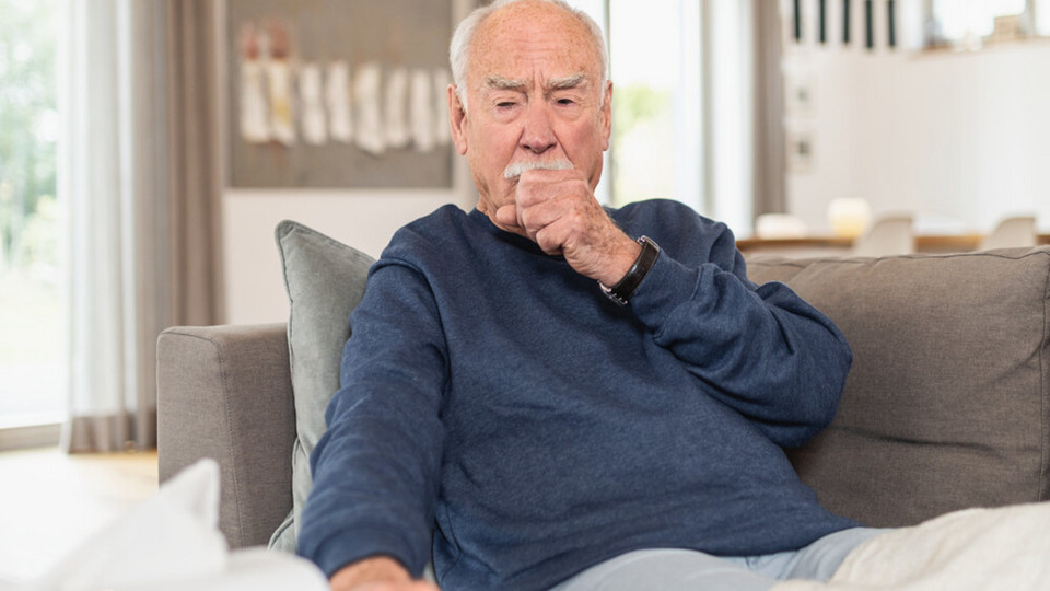Schleim im Hals lösen: So werden Sie den Frosch los Älterer Mann sitzt hustend mit Faust vor dem Mund auf Sofa