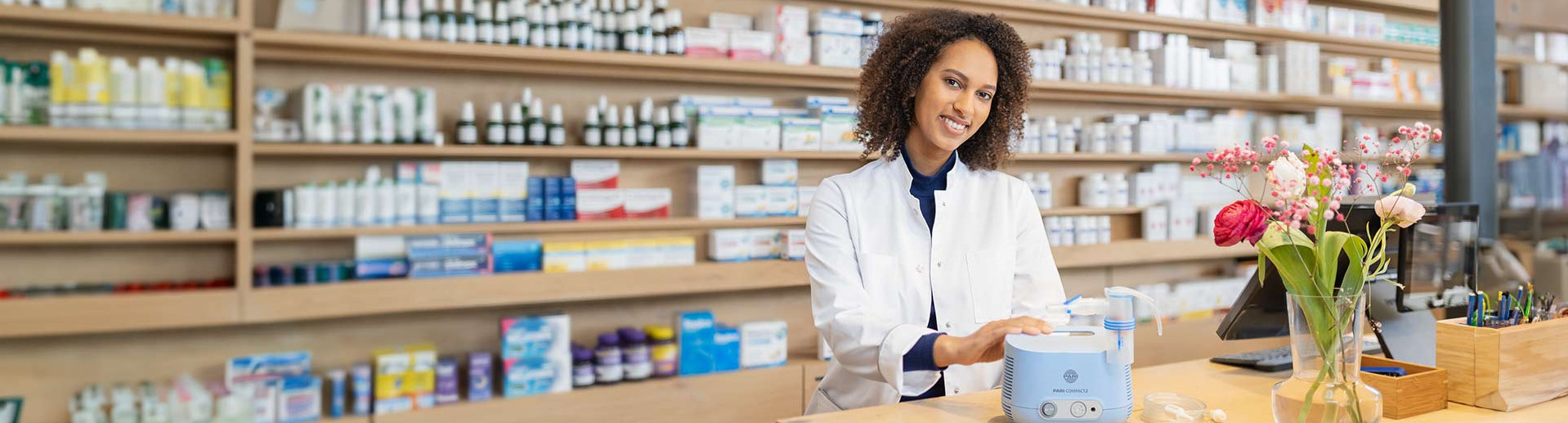 In einer Apotheke steht eine Apothekerin hinter der Theke und erklärt die Funktionsweise des PARI COMPACT2 Inhaliergerätes, das vor ihr auf der Theke steht. Lächelnd legt sie dabei die rechte Hand auf das Gerät. Rechts neben Ihr steht eine Vase mit Blumen. Die Apotheke ist modern, hell und freundlich.