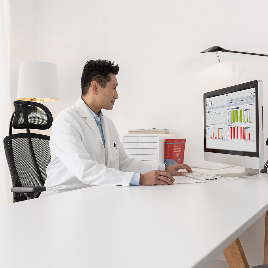 Arzt im weißen Kittel analysiert medizinische Daten am Computer in heller, moderner Büro-Umgebung.