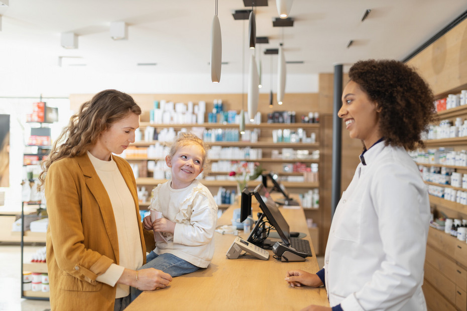 Junge Mutter, deren Tochter auf der Theke einer Apotheke vor ihr sitzt, im Gespräch mit Apothekerin