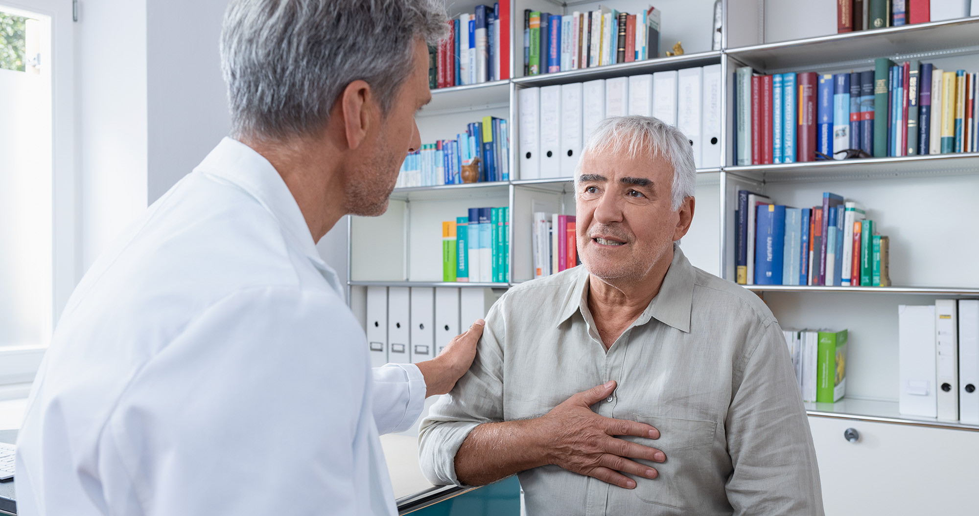 Ein älterer Mann sitzt in einer Arztpraxis, hält seine Hand auf die Brust und spricht mit einem Arzt im weißen Kittel, der ihm beruhigend eine Hand auf die Schulter legt.