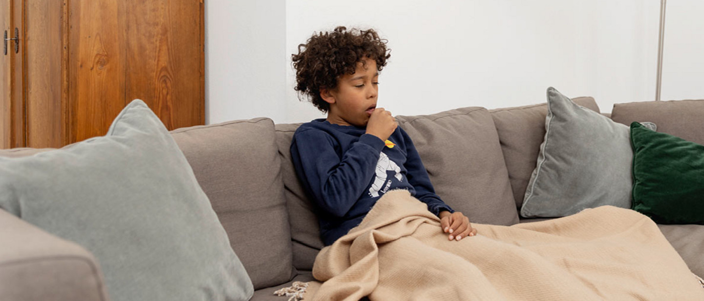 A sick boy sits at home on the sofa under a blanket and has to cough