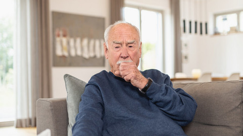 Elderly man sitting on the couch and coughing in his fist