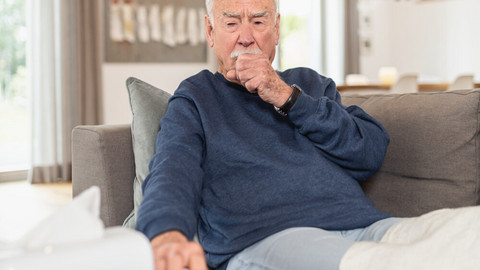 Husten mit Auswurf – Ursachen und Tipps gegen lästigen Schleim Älterer Mann sitzt hustend auf Sofa, eine Hand als Faust vor dem Mund, die andere Hand greift nach einem Taschentuch