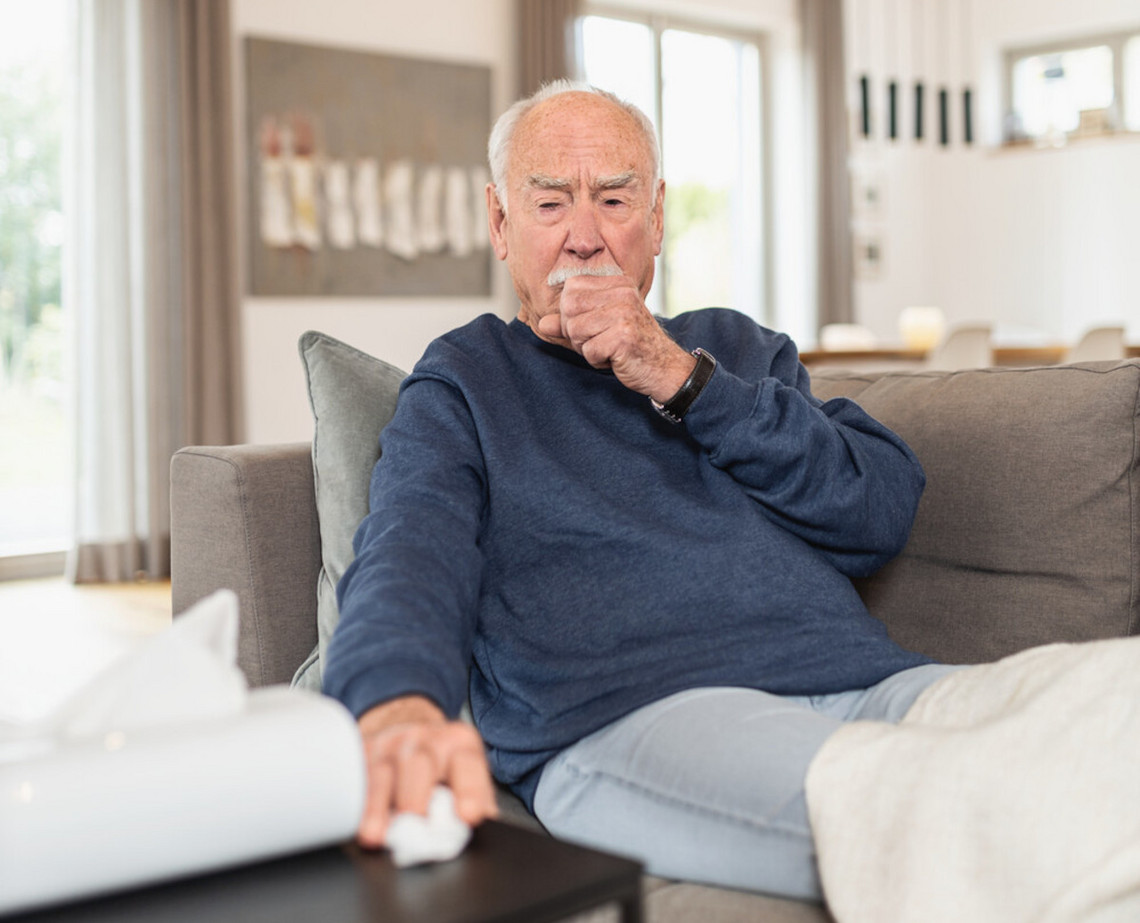 Elderly man sitting on sofa coughing, one hand as a fist in front of his mouth, the other hand reaching for a handkerchief
