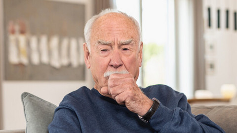 Älterer Mann sitzt hustend auf Sofa, eine Hand als Faust vor dem Mund, die andere Hand greift nach einem Taschentuch