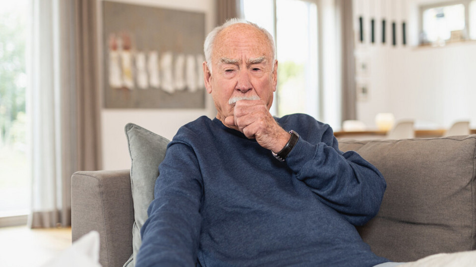 Älterer Mann sitzt hustend auf Sofa, eine Hand als Faust vor dem Mund, die andere Hand greift nach einem Taschentuch