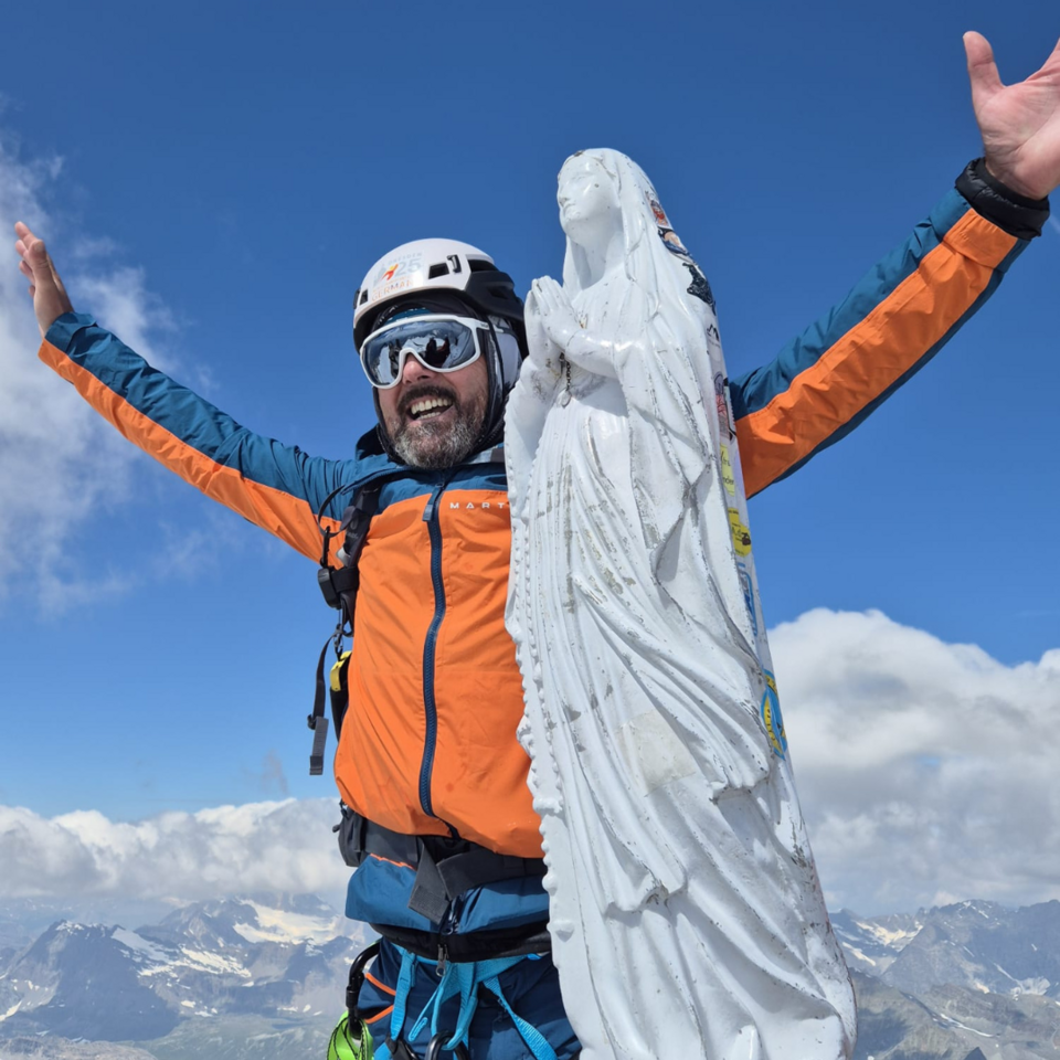 Ein lächelnder Mann mit Bergausrüstung steht neben der Madonna auf dem Gipfel des Gran Paradiso