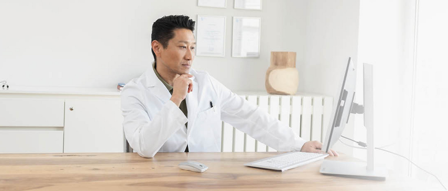 A doctor in a white coat is sitting at a desk, working intently on a computer in a bright office.