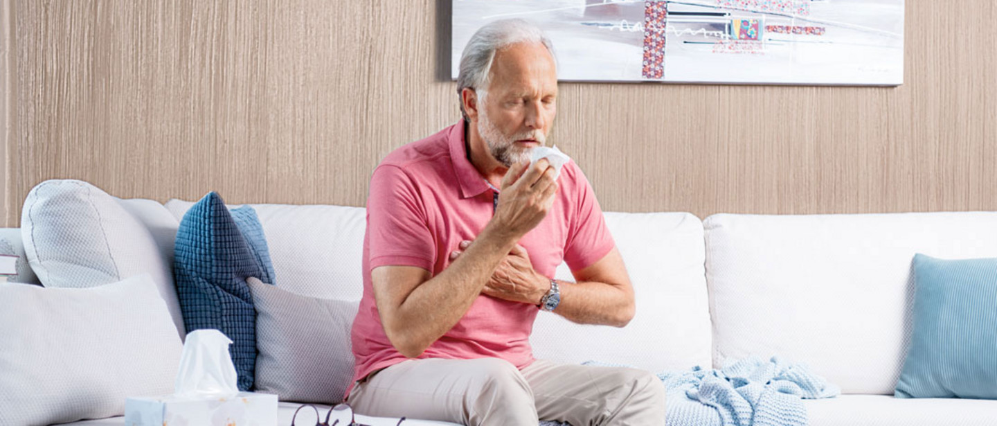 An elderly man sits on the sofa, coughing heavily and holding his hand in front of his chest