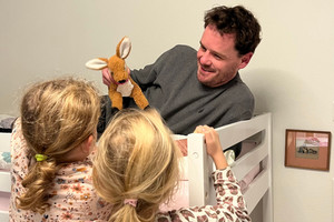 Father lies on bunk bed with cuddly toy in hand, in front of him his daughters look up to him