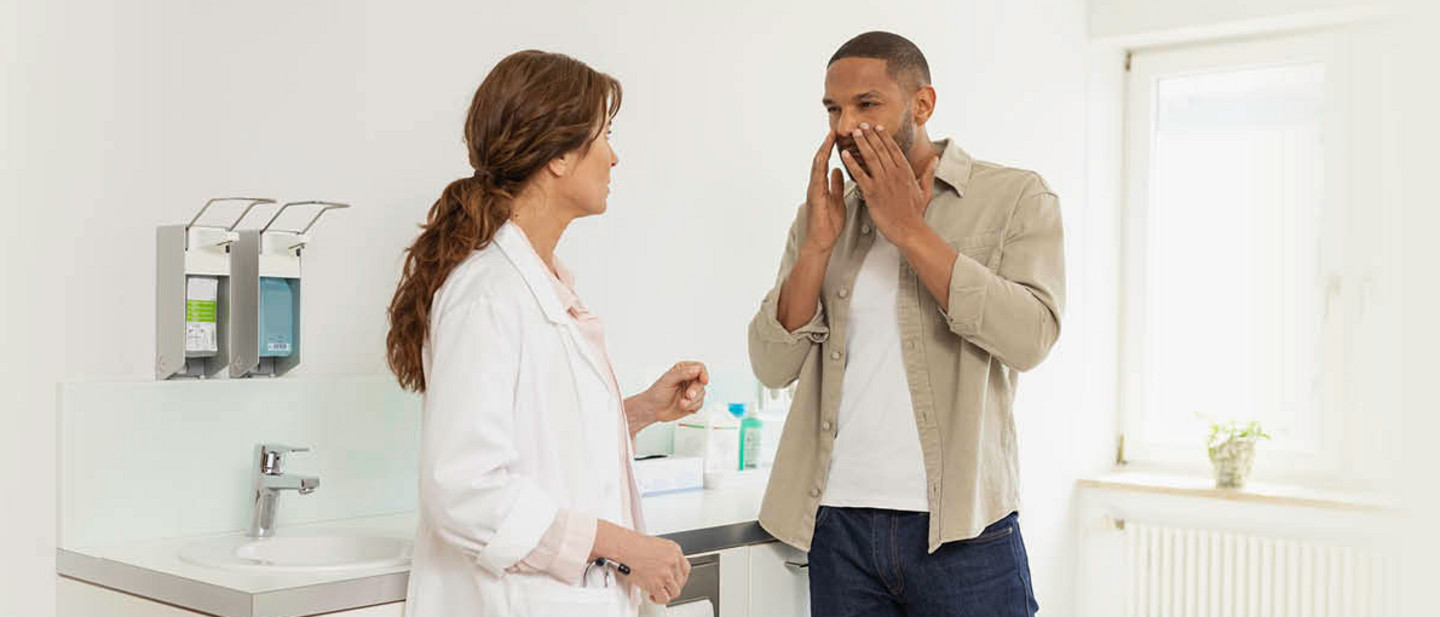 Arzt im weißen Kittel spricht mit Patient, der auf sein Gesicht zeigt – Szene in hellem Behandlungsraum.