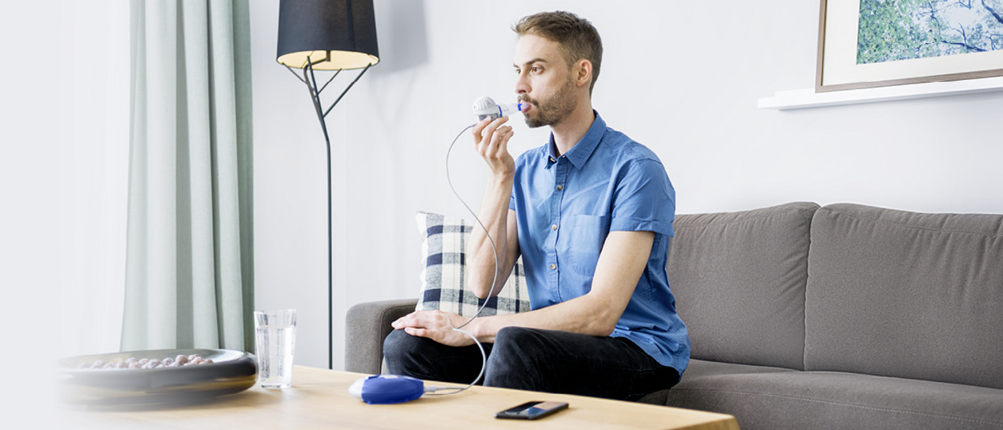 Ein jüngerer Mann im blauen Hemd sitzt im Wohnzimmer auf einem grauen Sofa und inhaliert mit einem Vernebler, der mit einem Controller auf dem Tisch verbunden ist.