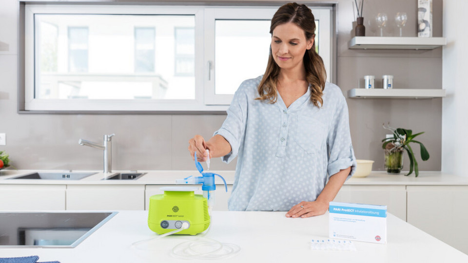 Junge Frau steht in Küche an Arbeitsfläche und füllt Inhalationslösung PARI Protect mit Ectoin in Inhalationsgerät PARI BOY Junior