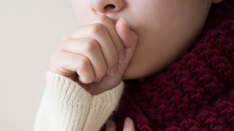 Frau hustet in die geschlossene Faust, die andere Hand hält sie sich an den Hals