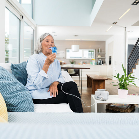 Elderly woman sits on sofa and inhales with inhalation device