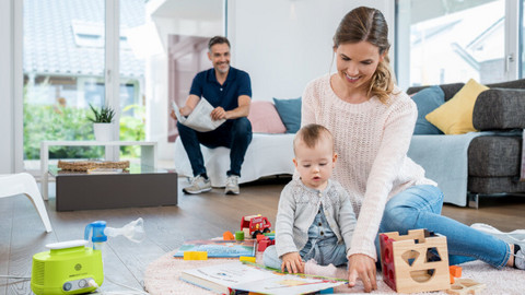 Mutter sitzt mit Kleinkind auf dem Teppich und spielt, daneben steht ein Inhalationsgerät, Vater sitzt im Hintergrund auf Sofa