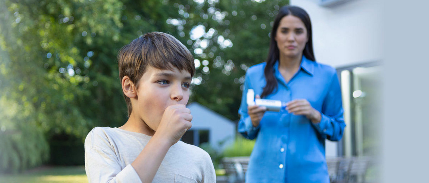 Junge mit braunen Haaren steht in einem Garten und hält sich die Hand vor den Mund und hustet. Im Hintergrund steht seine Mutter mit langen dunklen Haaren in einem blauen Hemd, die eine VORTEX Inhalierhilfe in den Händen hält und besorgt schaut. Das Bild vermittelt eine Szene, in der Asthma oder Atembeschwerden behandelt werden.