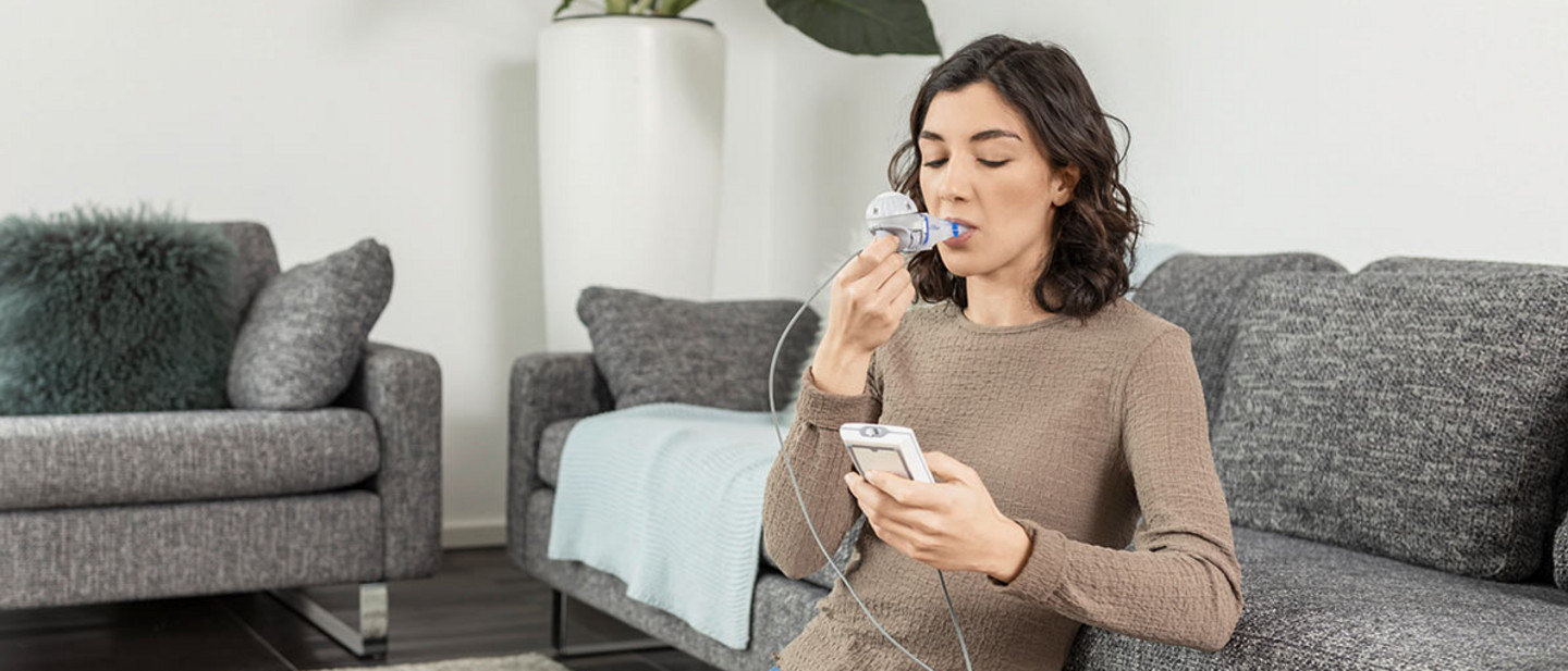 Eine junge Frau mit dunklen Haaren sitzt in einem modern eingerichteten Wohnzimmer vor einem Sofa. Sie hält ein Inhalationsgerät in der Hand und inhaliert durch ein Mundstück, während sie auf den dazugehörigen Controller schaut.