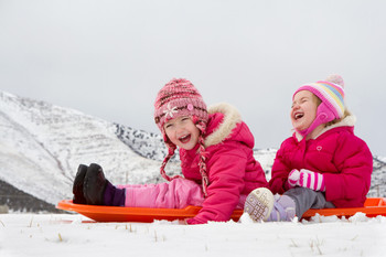 Zwei kleine Kinder sitzen im Schnee auf ihren Schlitten und lachen