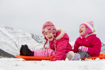 Zwei kleine Kinder sitzen im Schnee auf ihren Schlitten und lachen