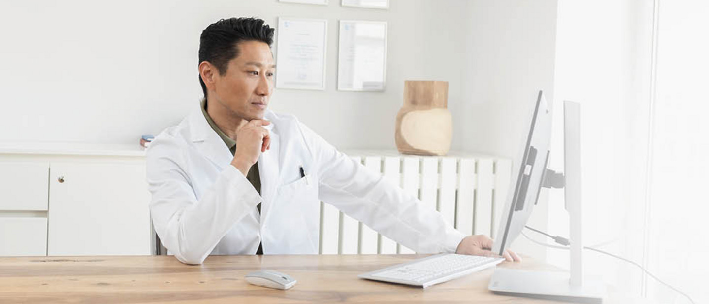 一位身穿白袍的醫生坐在書桌前，在明亮的辦公室裡專注地操作電腦。