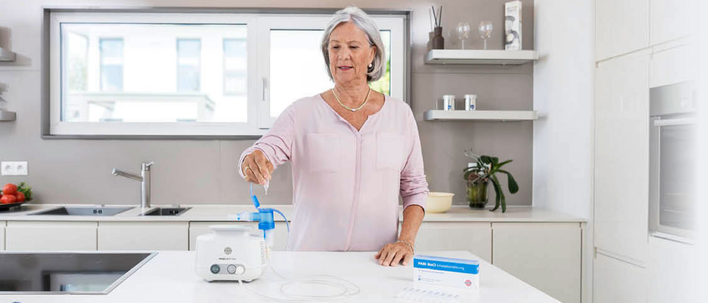 Ein Frau nutzt einen PARI-Inhalator in einer moderner Küche zur Atemtherapie, Medikamentenampullen und Zubehör liegen bereit.