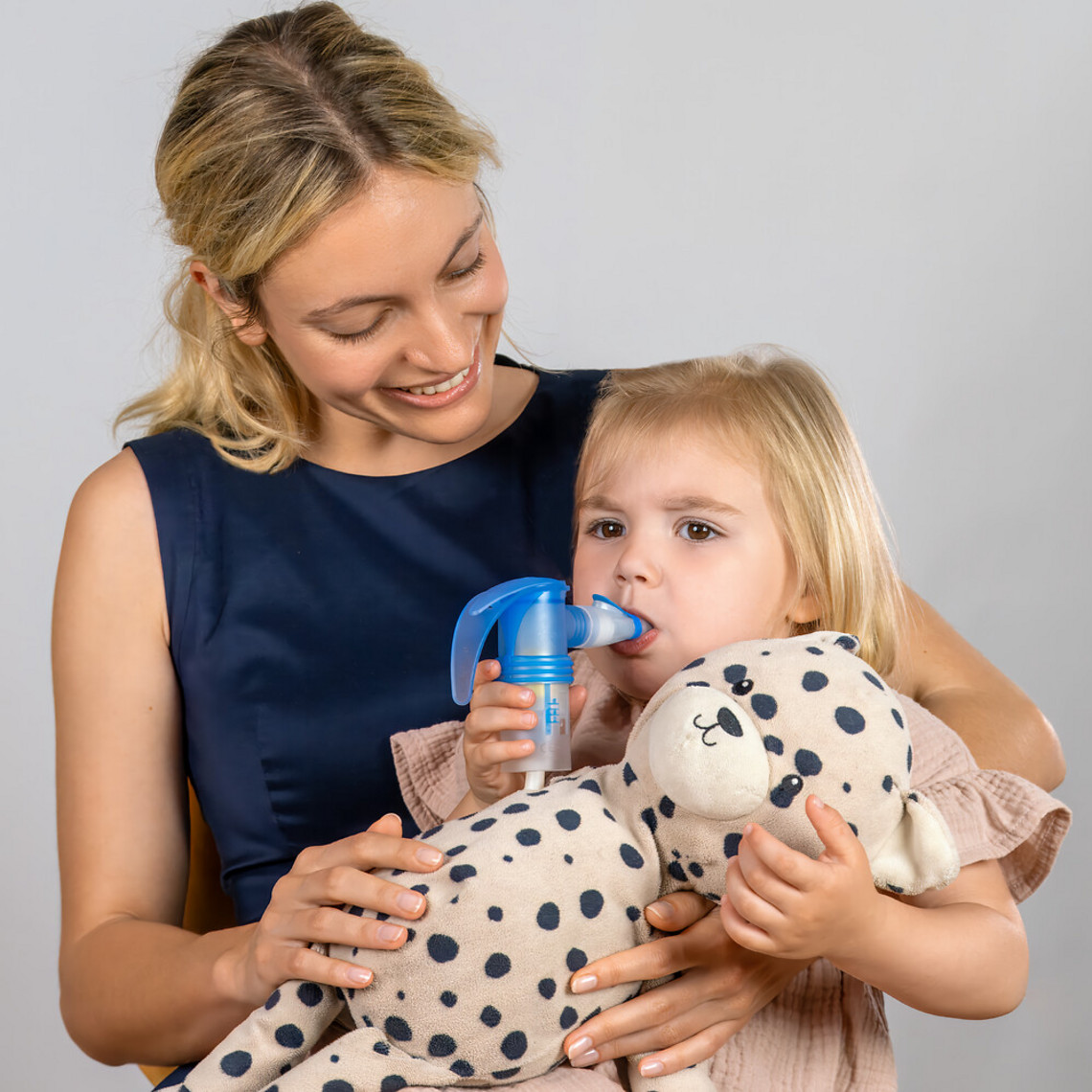 Tochter sitzt bei Mutter auf dem Schoß, sie inhaliert mit Vernebler und hält ein Kuscheltier