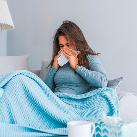 Junge Frau sitzt mit Decke auf dem Sofa und schnäuzt in ein Taschentuch