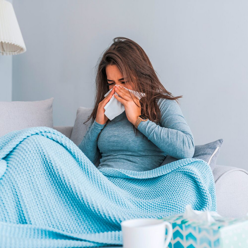 Junge Frau sitzt mit Decke auf dem Sofa und schnäuzt in ein Taschentuch