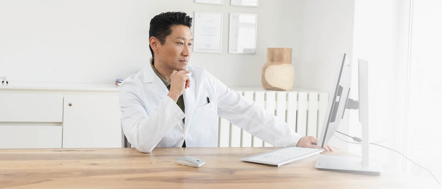 Ein Arzt im weißen Kittel sitzt am Schreibtisch und arbeitet konzentriert am Computer in einem hellen Büro.