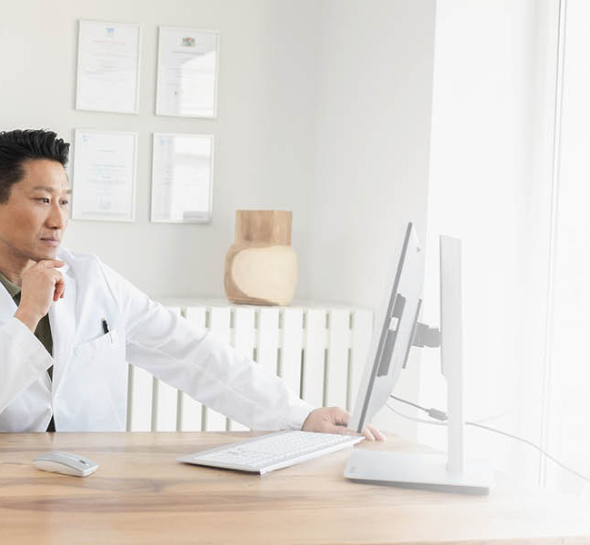 Ein Arzt im weißen Kittel sitzt am Schreibtisch und arbeitet konzentriert am Computer in einem hellen Büro.