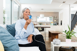 Elderly woman sits on sofa and inhales with inhalation device